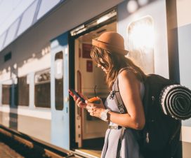 Railroad theme. Beautiful young woman with a backpack uses the phone while standing near the railroad train on the platform. Cheap travel