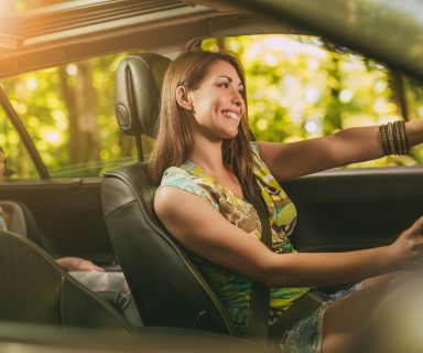 Young beautiful smiling woman driving a car. Her cute daughter sitting on rear and enjoying.